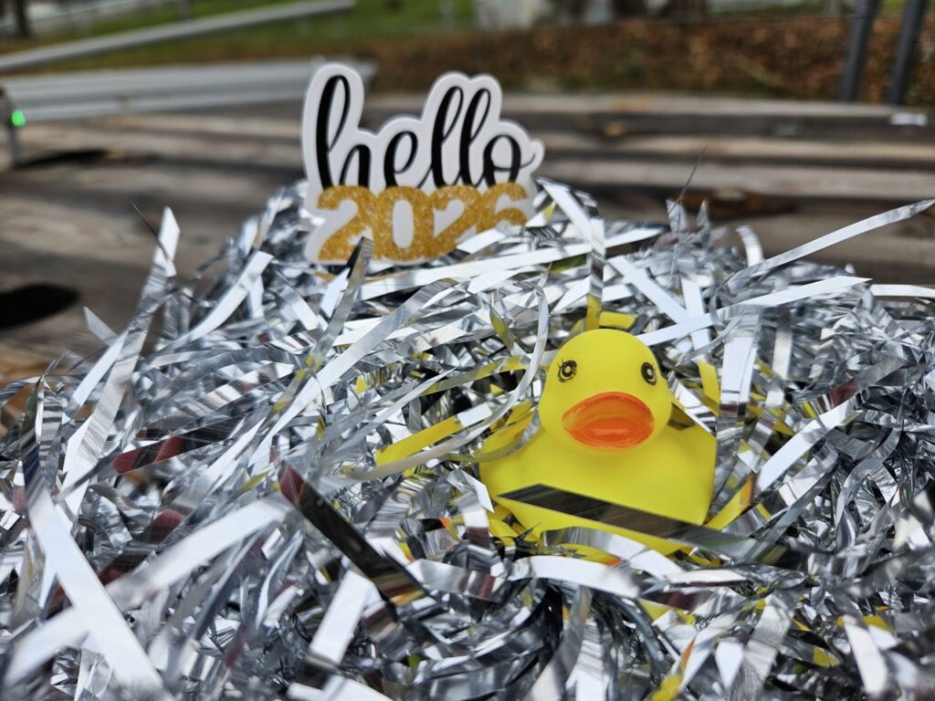 Gelbe Quietscheente sitzt in silbernem Lametta im Freien, darüber ein Schriftzug „hello 2026“, unscharfer Hintergrund mit Straße und Grünfläche